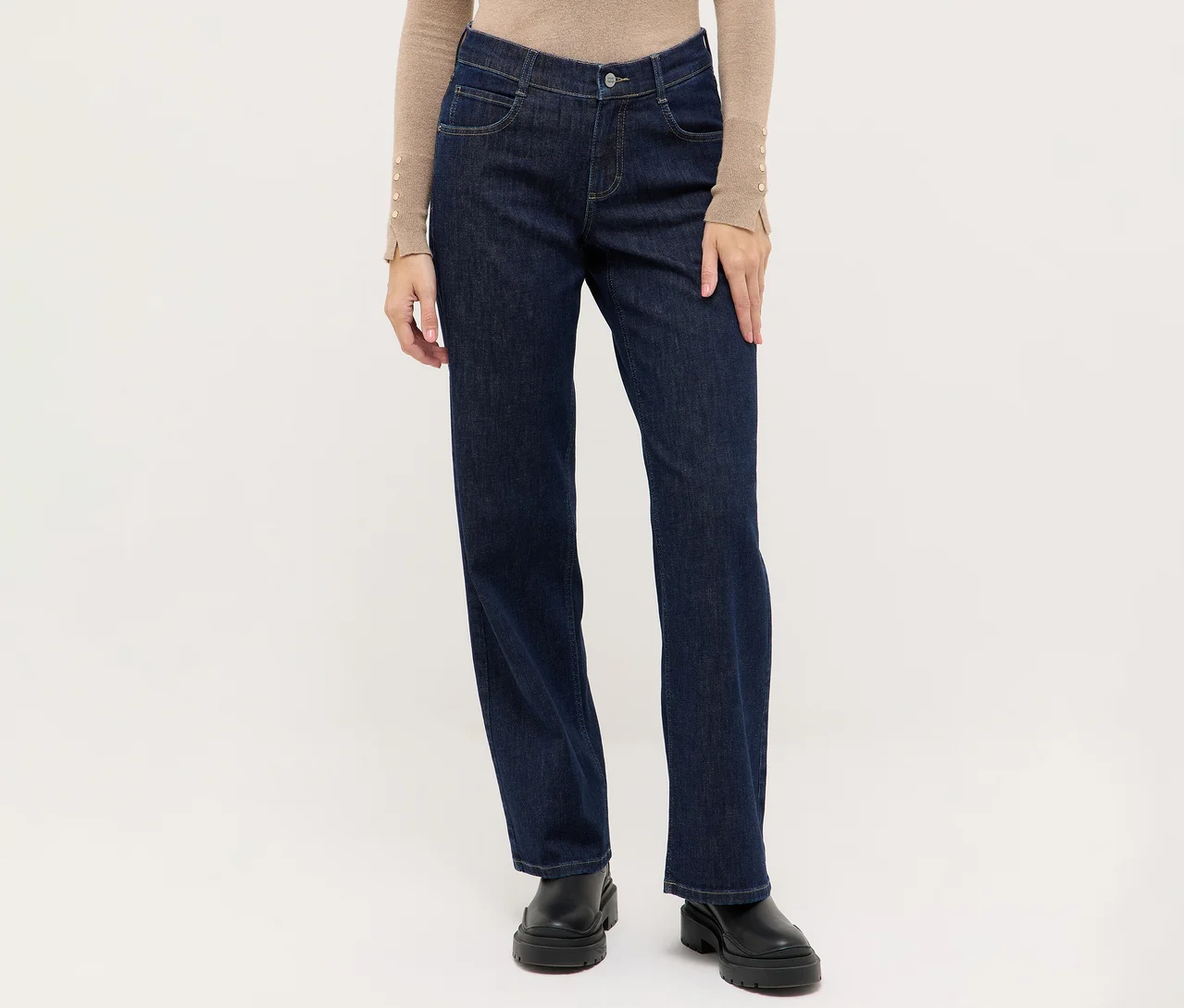 Person trägt dunkelblaue Jeans mit geradem Bein, ein beiges Langarmshirt mit Knopfdetails an den Ärmelbündchen und schwarze klobige Stiefel.