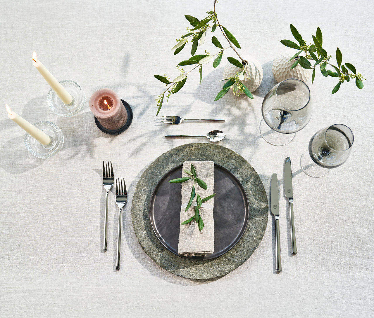 Elegant gedeckter Tisch für zwei Personen mit Besteck aus dem ZWILLING »Aberdeen« Besteck-Set. Auf hellem Tischtuch: dunkler Teller auf grauem Unterteller, Leinen-Serviette mit Olivenzweig, zwei Gabeln, zwei Messer, Gabel und Löffel. Rechts zwei graue Weingläser, links drei Kerzen, oben zwei Vasen mit Olivenzweigen.