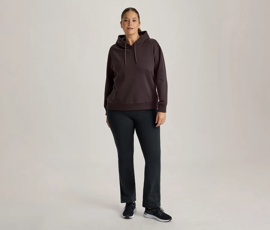 Eine Frau steht frontal und trägt einen dunkelbraunen Kapuzenpullover, eine dunkle Hose und schwarze Turnschuhe.