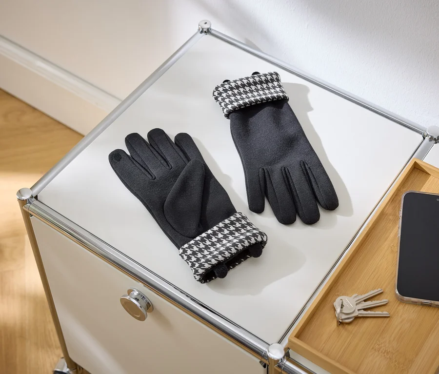 Ein Paar schwarze Handschuhe mit Hahnentrittmuster-Bündchen liegt auf einer weißen Oberfläche. Daneben befinden sich ein Smartphone und Schlüssel auf einem Holztablett.