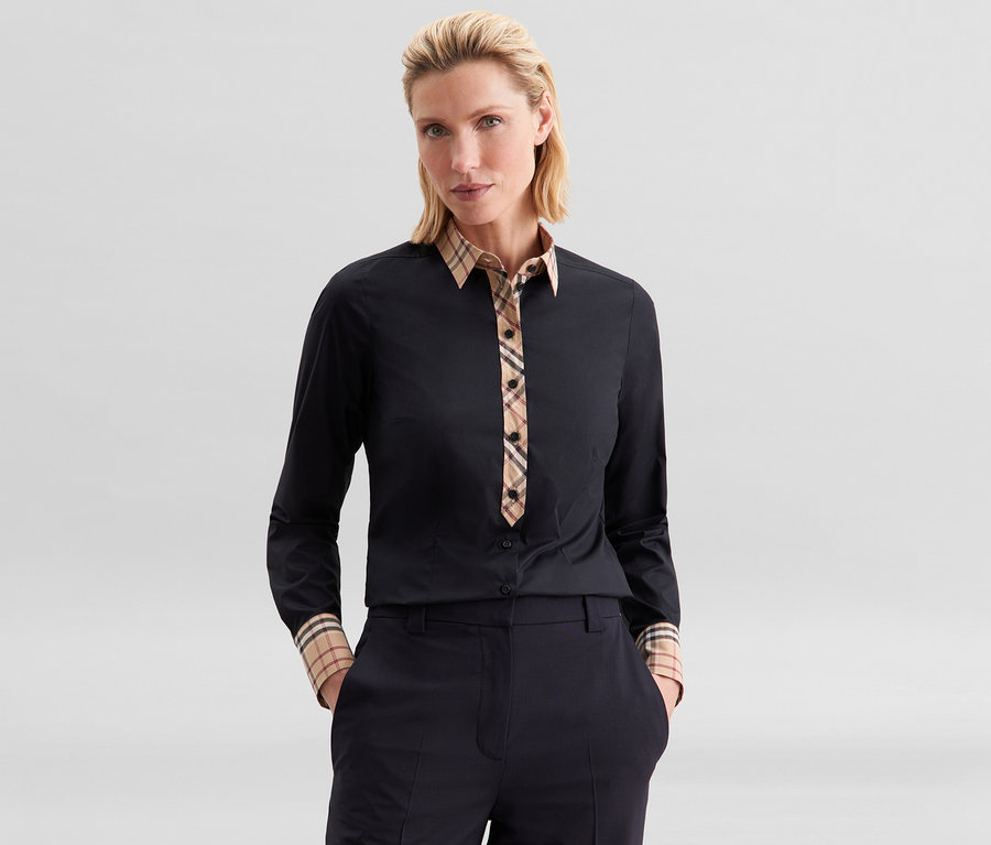 Eine Frau trägt eine schwarze Seidensticker Popeline-Bluse mit Karodetail an Kragen, Knopfleiste und Manschetten sowie eine dunkle Hose.