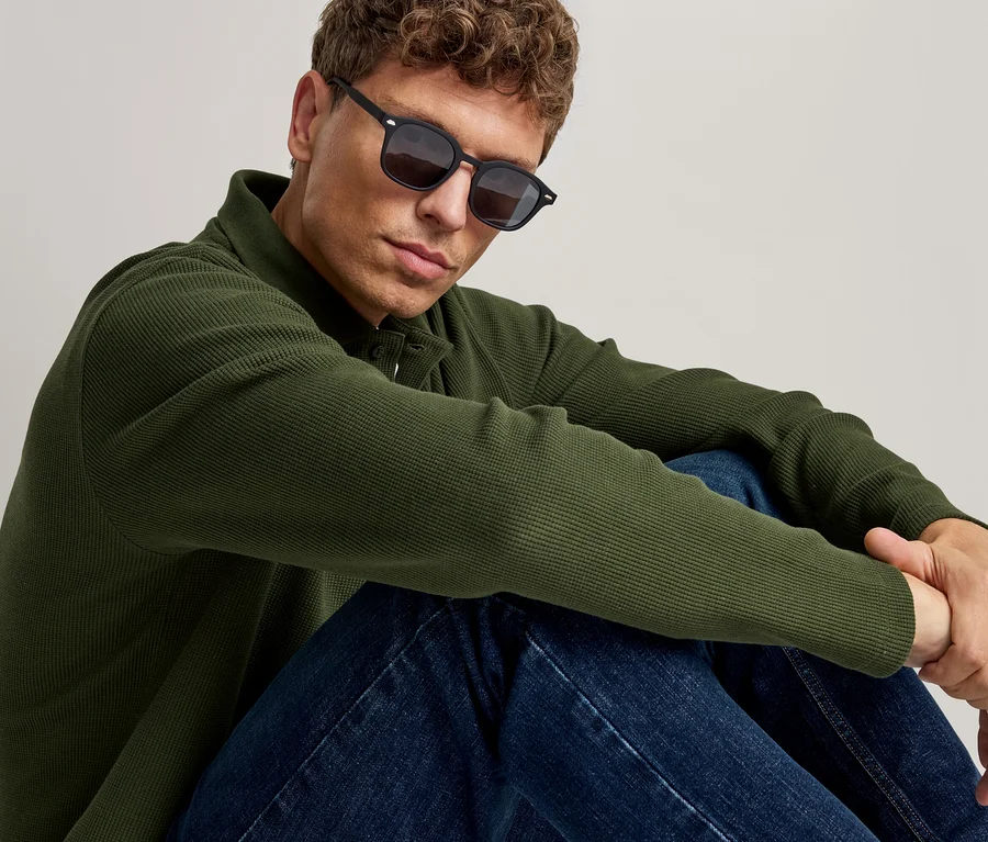 Mann mit schwarzer Sonnenbrille, grünem Langarm-Poloshirt und blauen Jeans, sitzend und nach vorne blickend.