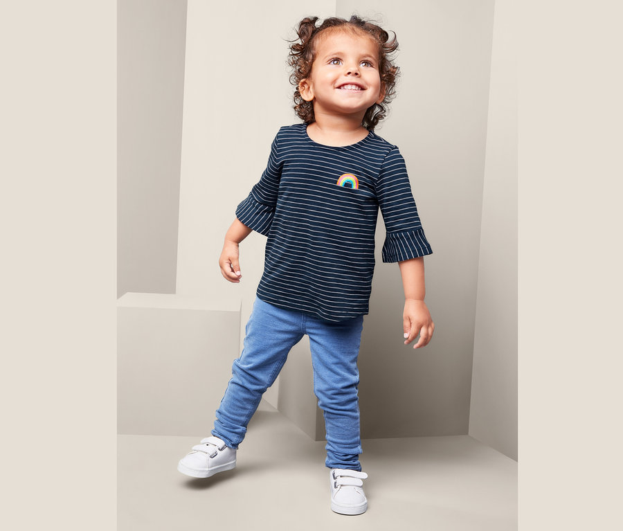Ein Mädchen trägt ein Streifen-Shirt mit Regenbogen, blaue Jeans und weiße Schuhe.