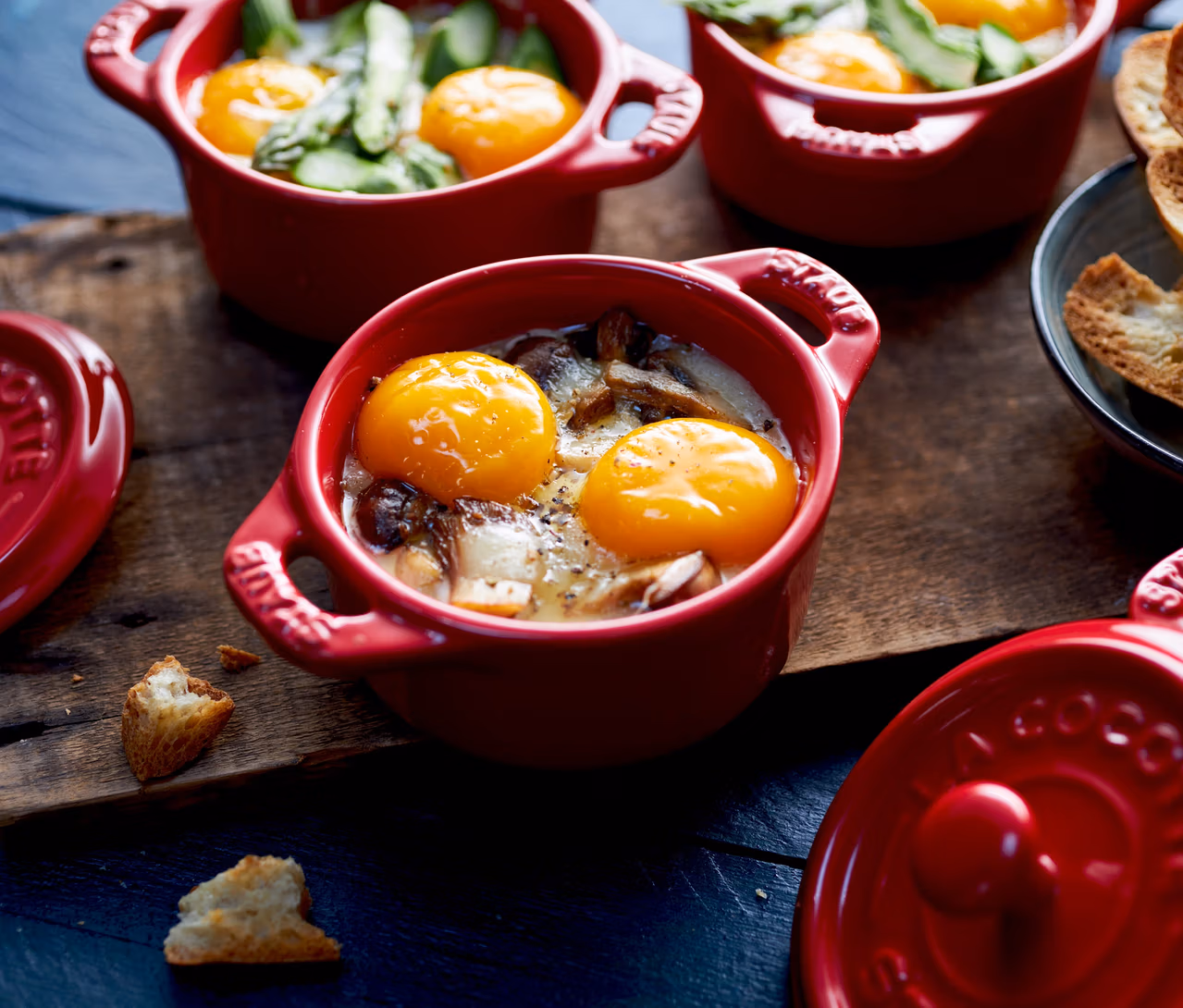 Drei kirschrote STAUB Keramik-Cocottes mit gebackenen Eiern, Pilzen und Spargel stehen auf einem Holzbrett. Zwei passende Deckel und geröstete Brotscheiben liegen daneben.
