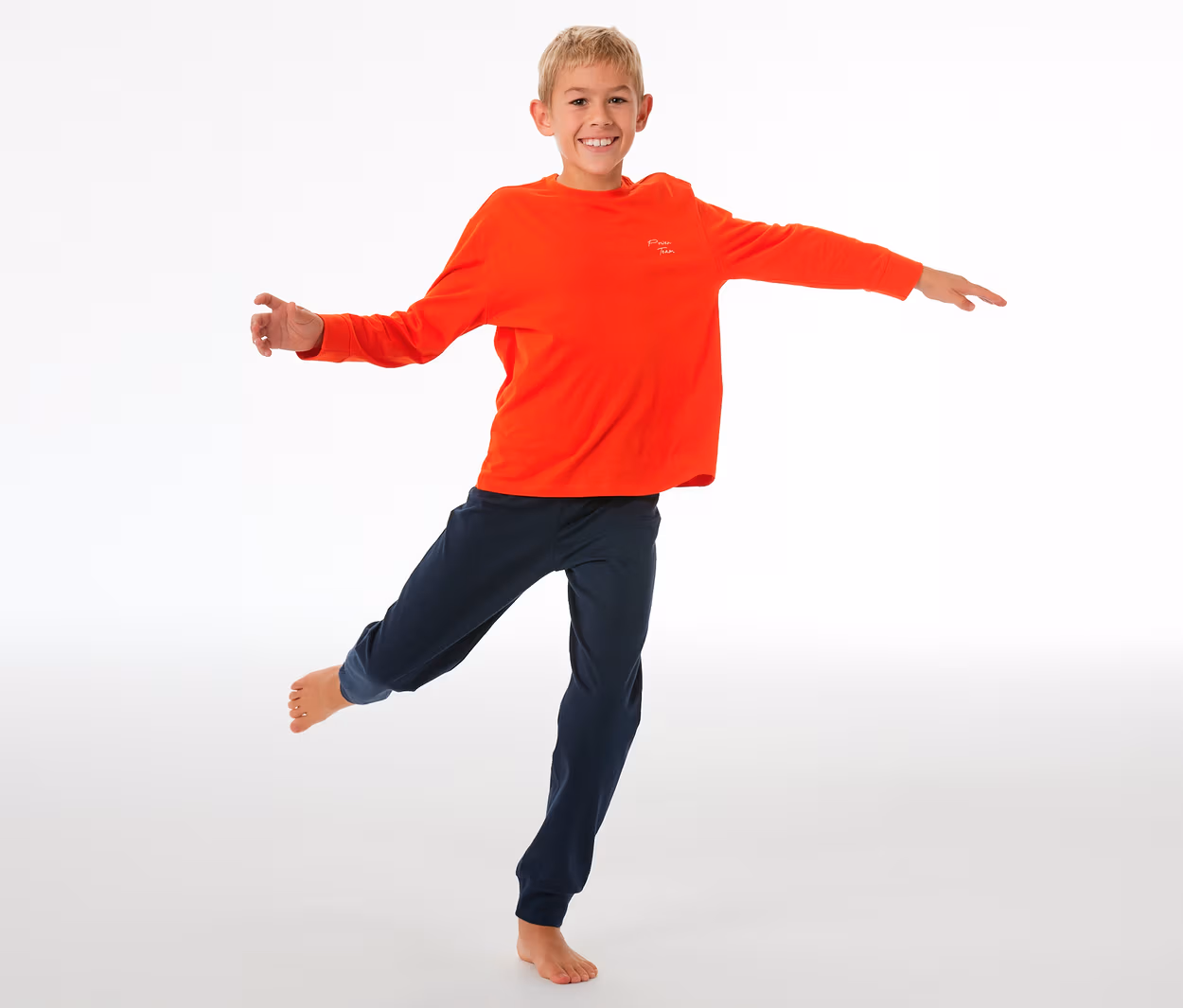 Lächelnder Junge balanciert auf einem Bein, trägt einen SCHIESSER Kinder-Pyjama mit Bündchen: orangefarbenes Langarmshirt und dunkelblaue Hose.