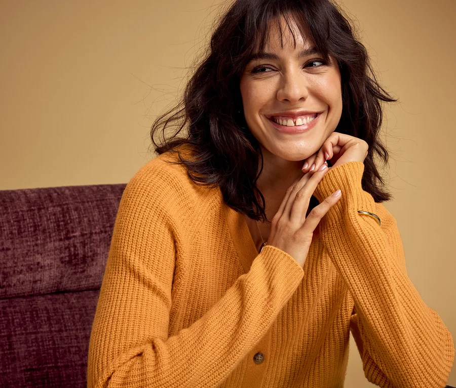 Lächelnde Frau mit dunklen Haaren in einem orangefarbenen Strickcardigan und einem goldenen Armband, die ihre Hände unter dem Kinn auf einem lila Stuhl abstützt.