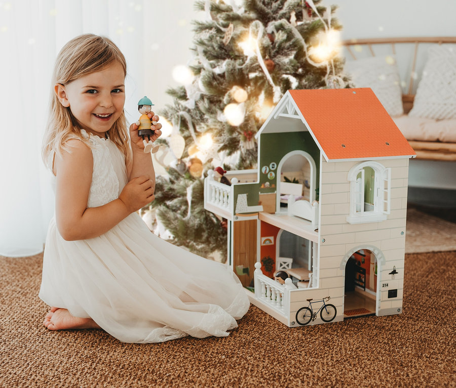 Lächelndes Mädchen im Kleid kniet neben einem Small foot Puppenhaus mit Dachterrasse und hält eine Figur.