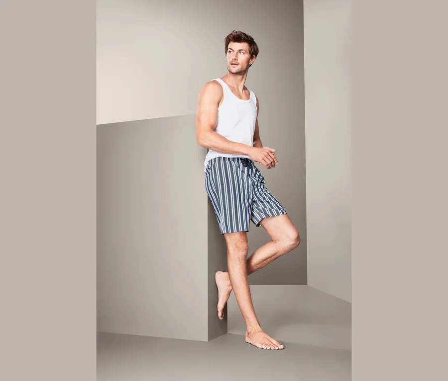 Mann lehnt an Wand, trägt weißes Tanktop und Web-Pyjamashorts.
