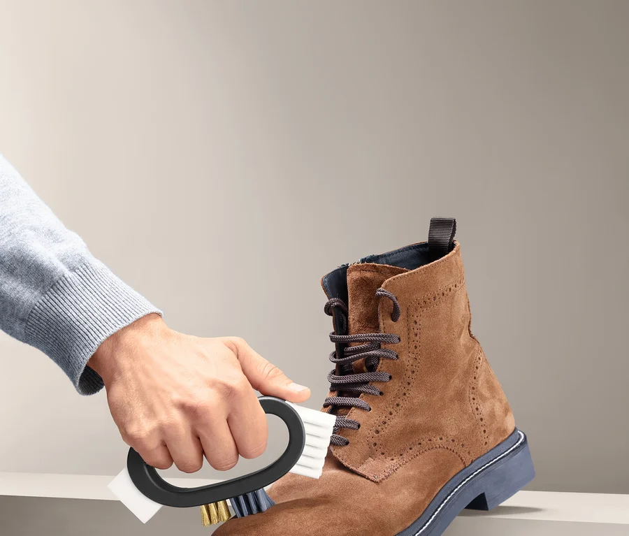 Eine Hand reinigt einen braunen Wildlederstiefel mit einer 3-in-1-Universal-Schuhbürste.