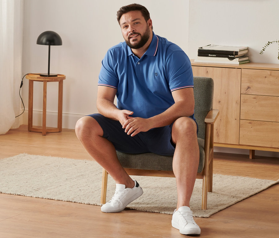 Mann sitzt im Sessel und trägt eine marineblaue Chino-Shorts, ein royalblaues Piqué-Poloshirt und Sneaker.