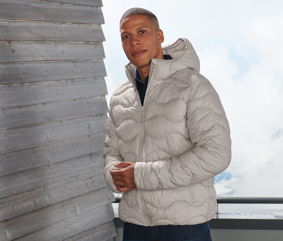 Mann posiert auf einem Balkon in beiger Steppjacke.