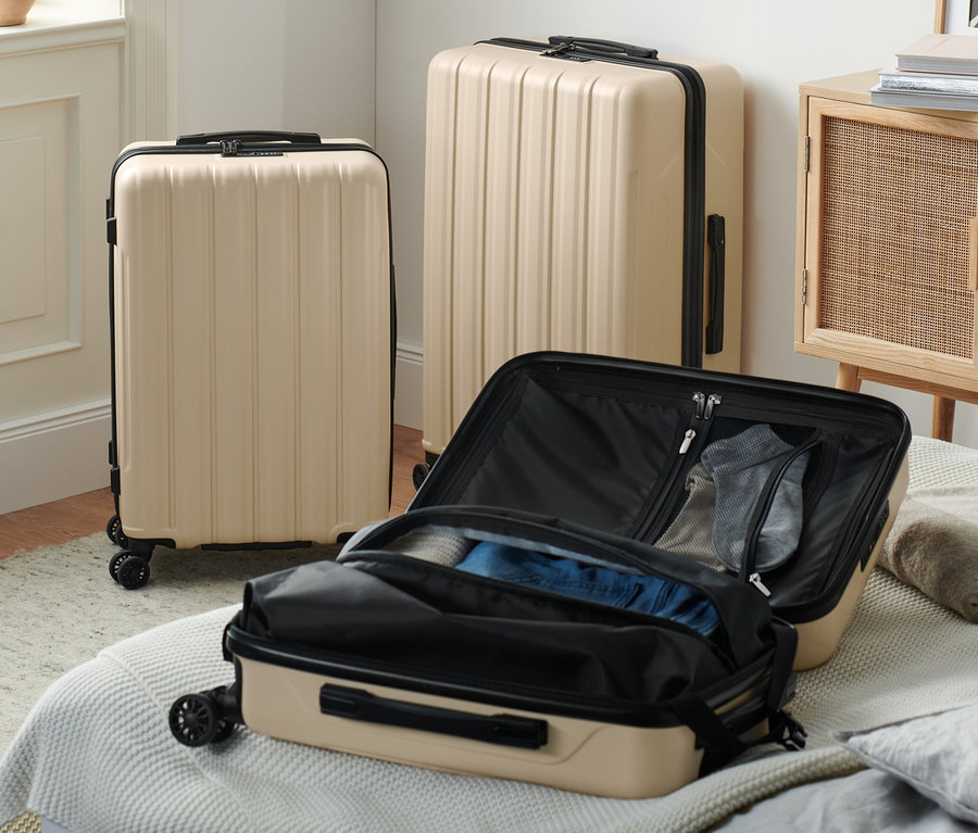 Drei beige Hartschalen-Koffer mit recyceltem Material stehen in einem Innenraum, einer ist auf einem Bett geöffnet.