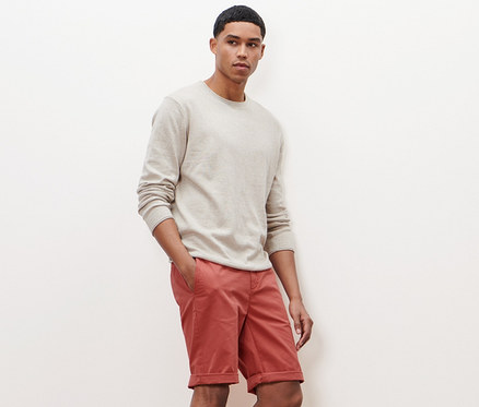 Mann posiert in beigem Pullover und Terracotta-Chino-Shorts.