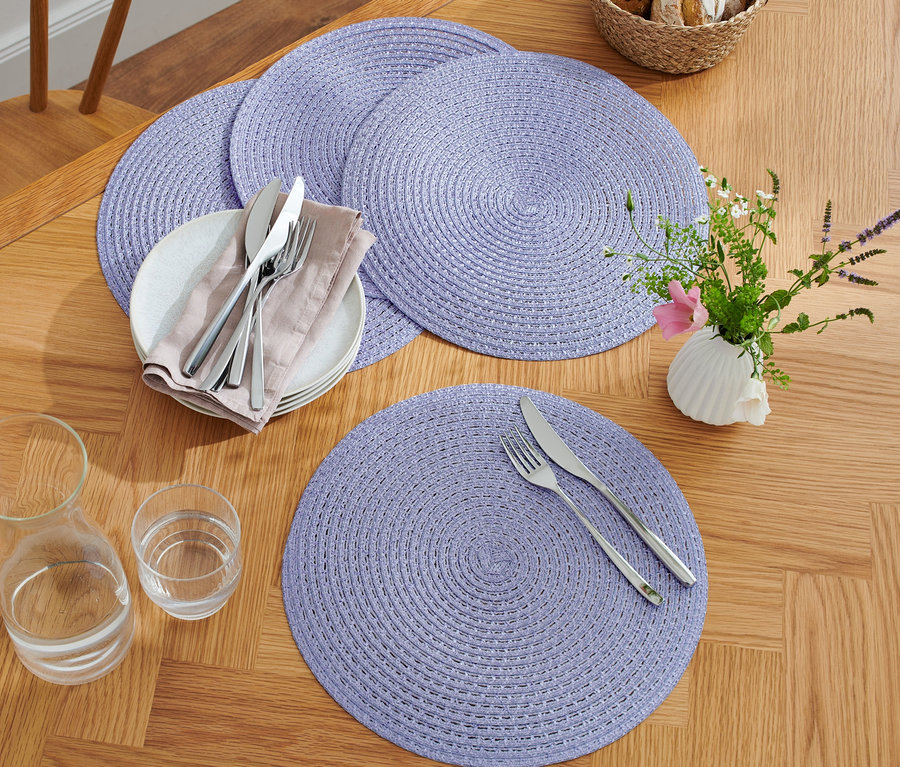 Vier lila Platzsets auf einem Holztisch mit Tellern, Besteck, Gläsern und einer Vase mit Blumen.