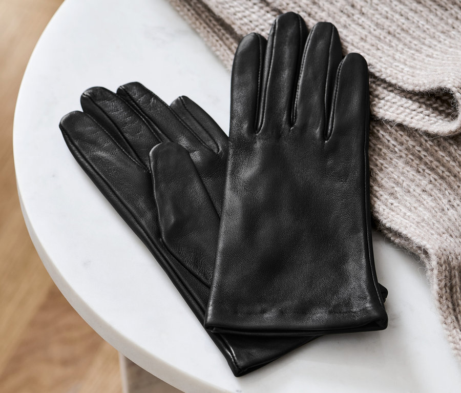Schwarze Lederhandschuhe liegen auf einem weißen Marmortisch neben einem beigen Strickpullover.