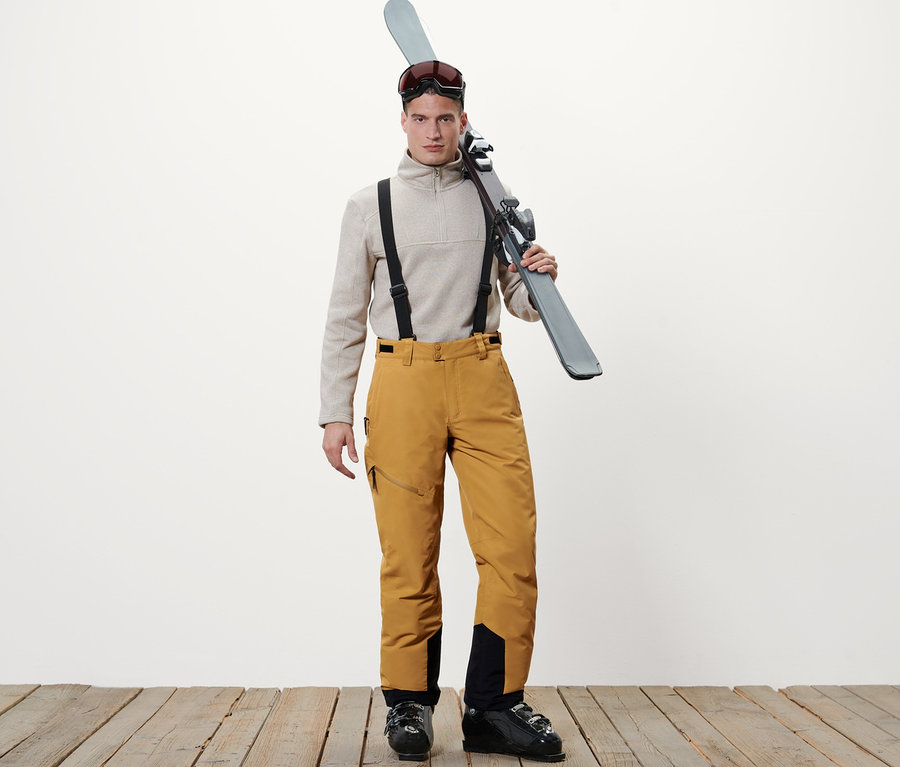 Mann steht mit Skiern über der Schulter, trägt einen beigen Pullover und eine Skihose in Camel.