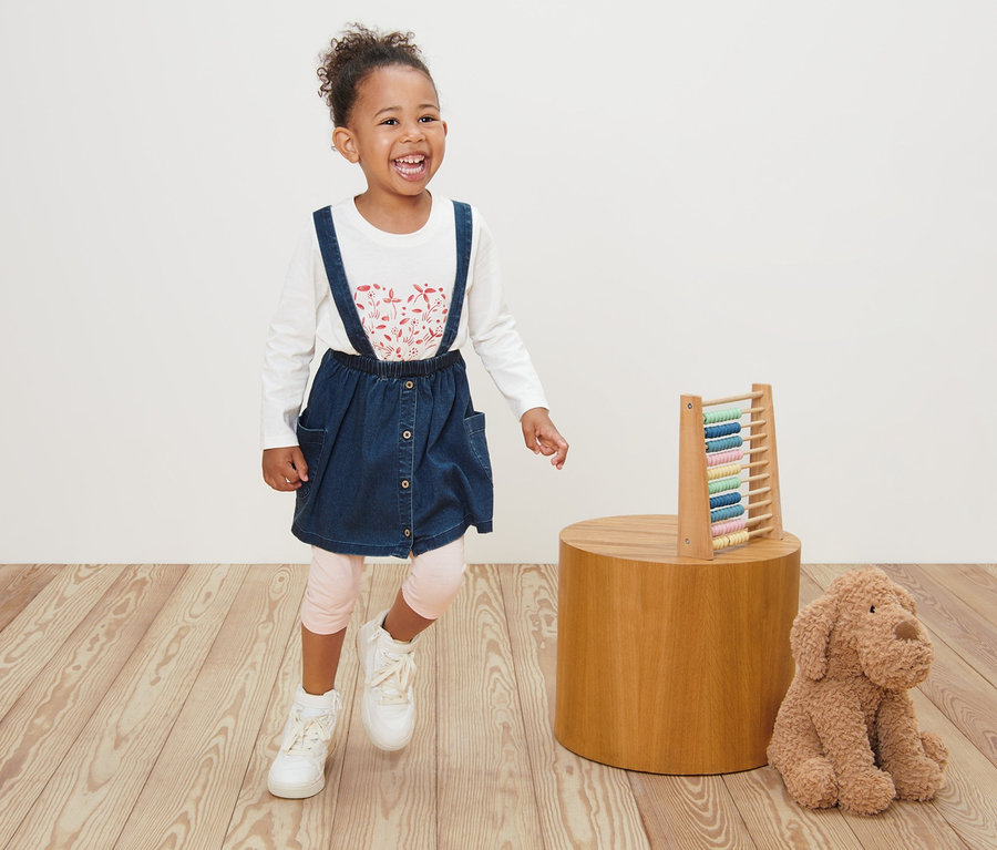 Lächelndes kleines Mädchen in Kleinkind-Denimrock und langärmeligem, weißen Oberteil steht neben einem Spielzeug auf einem Holzboden.