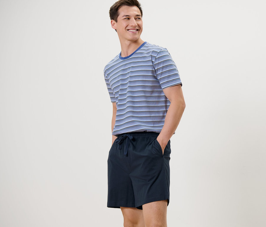 Mann steht und lächelt, er trägt einen blau-dunkelblauen Shorty-Pyjama.