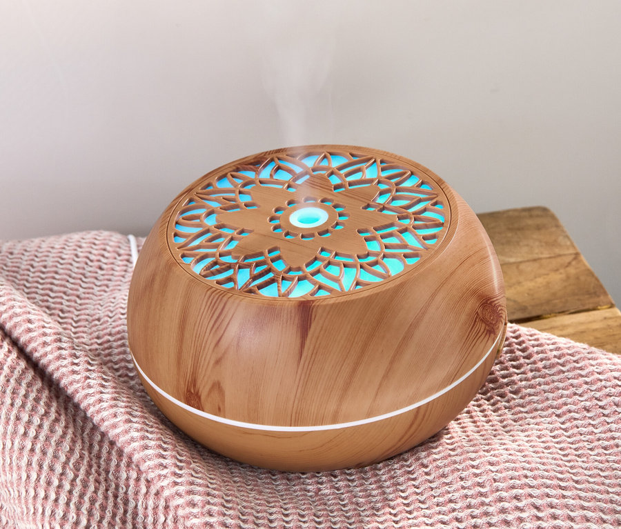 Ein Aroma-Diffusor mit Bluetooth®-Lautsprecher steht auf einer rosa Decke, aus dem Dampf aufsteigt.