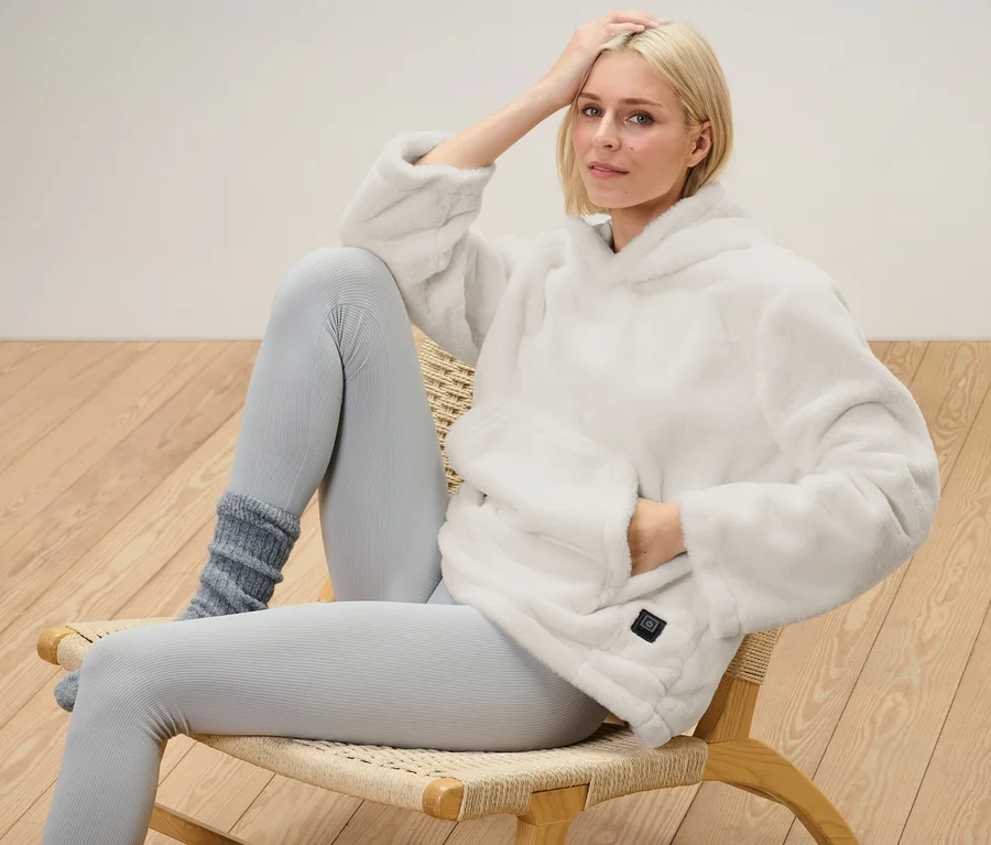 Blonde Frau sitzt auf einem Stuhl und trägt einen Akku-Heizpullover.