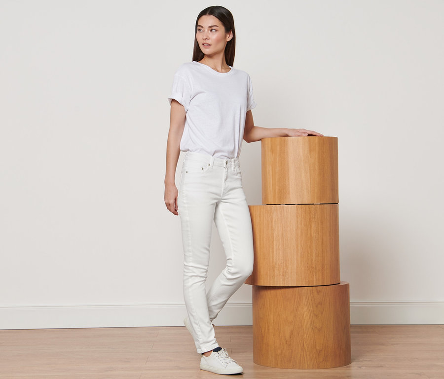 Eine Frau steht und lehnt sich an einen Stapel aus drei Holzsockeln. Sie trägt ein weißes T-Shirt und cremeweiße Colored Denim »Fit Emma«.
