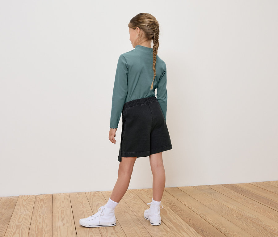 Mädchen mit Zopf trägt ein grünes Langarmshirt, schwarze Shorts und weiße Turnschuhe, von hinten gesehen.