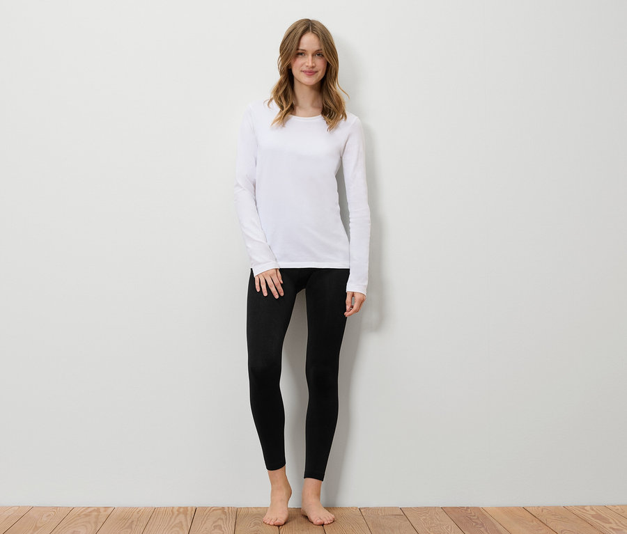 Frau steht vor einer weißen Wand, trägt ein weißes Langarmshirt und schwarze Leggings.
