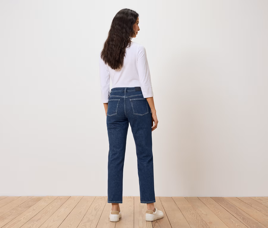 Eine Frau mit dunklen Haaren steht mit dem Rücken zur Kamera und trägt blaue Jeans und ein weißes T-Shirt.