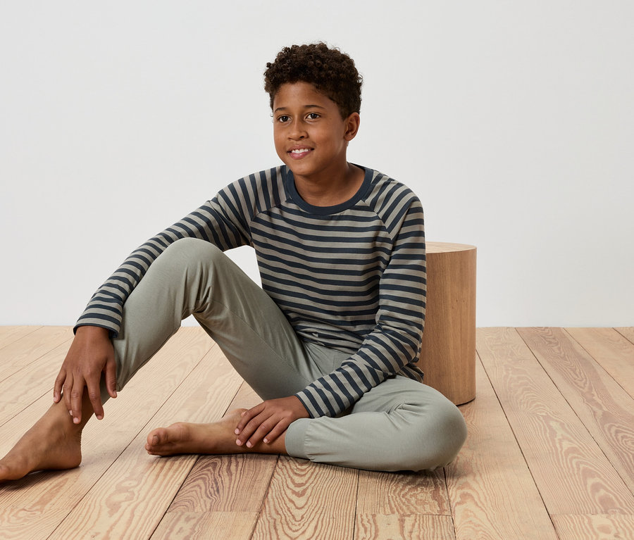 Ein junger Junge sitzt auf einem Holzboden, lehnt sich an einen kleinen Holztisch, trägt ein gestreiftes T-Shirt und eine Hose.