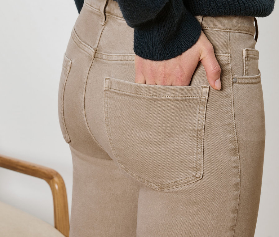 Detailaufnahme einer Frau, die eine braune Hose trägt und ihre Hand in die Hosentasche steckt. Anschnitt eines Sessels sichtbar.