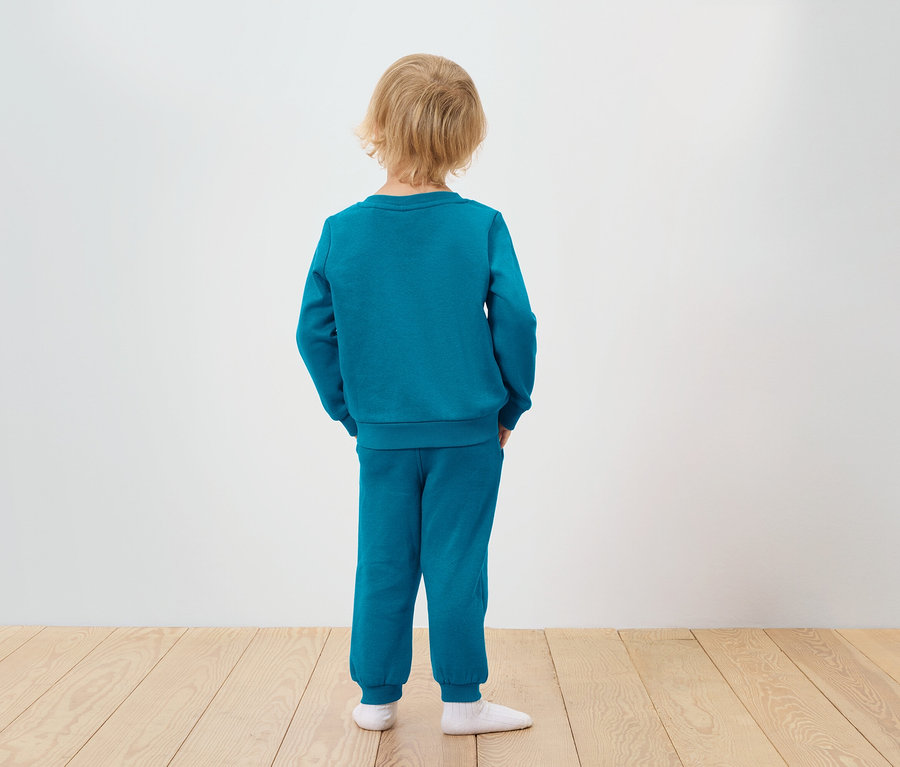 Junge im türkisfarbenen Trainingsanzug steht mit dem Rücken zur Kamera.