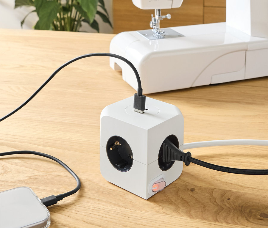 Auf einem Holztisch steht ein Steckdosenwürfel mit USB, an den ein Telefon und eine Nähmaschine angeschlossen sind.