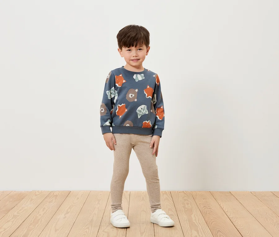 Junge trägt ein blaues Sweatshirt mit Bären- und Fuchs Muster, dazu beige Leggings und weiße Turnschuhe, steht auf einem Holzboden vor einer weißen Wand.
