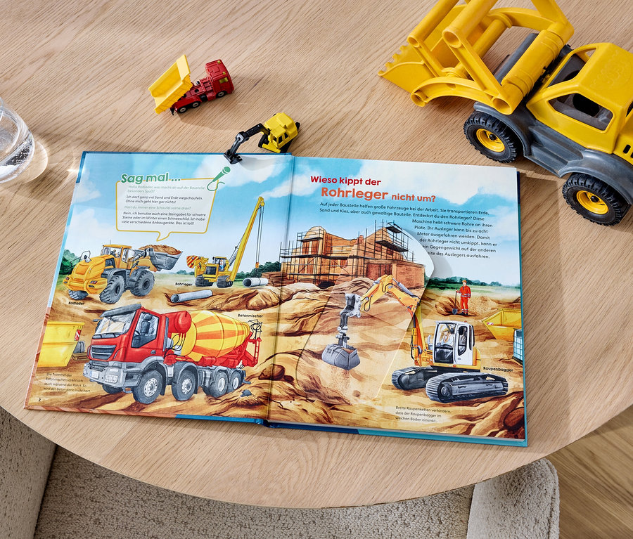 Offenes Buch mit Illustrationen von Baumaschinen und einer Baustelle, ergänzt durch Spielzeug-Lastwagen und ein Glas Wasser auf einem Holztisch.