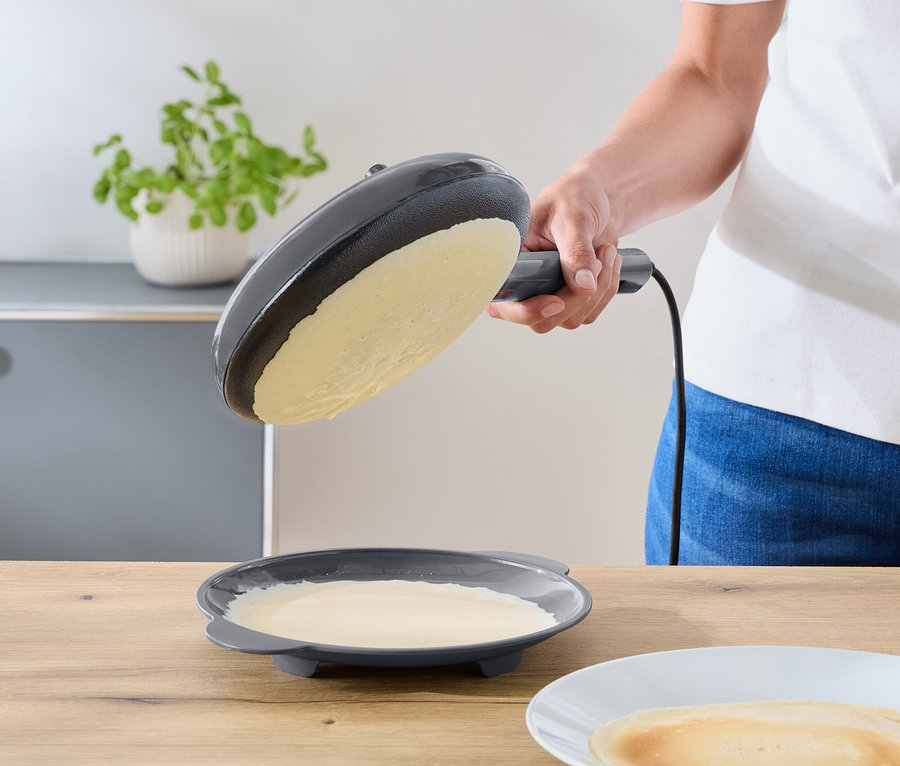 Eine Person neigt eine dunkelgraue elektrische Bratpfanne über einen Teller mit restlichem Teig. Ein fertiger Crêpe liegt auf einem weißen Teller auf einer Holzablage.