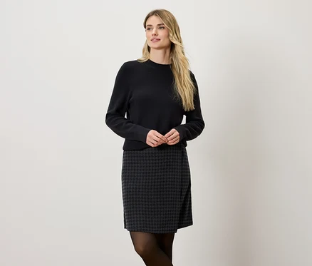Blonde Frau trägt einen schwarzen Pullover und einen grau-schwarzen Rock.