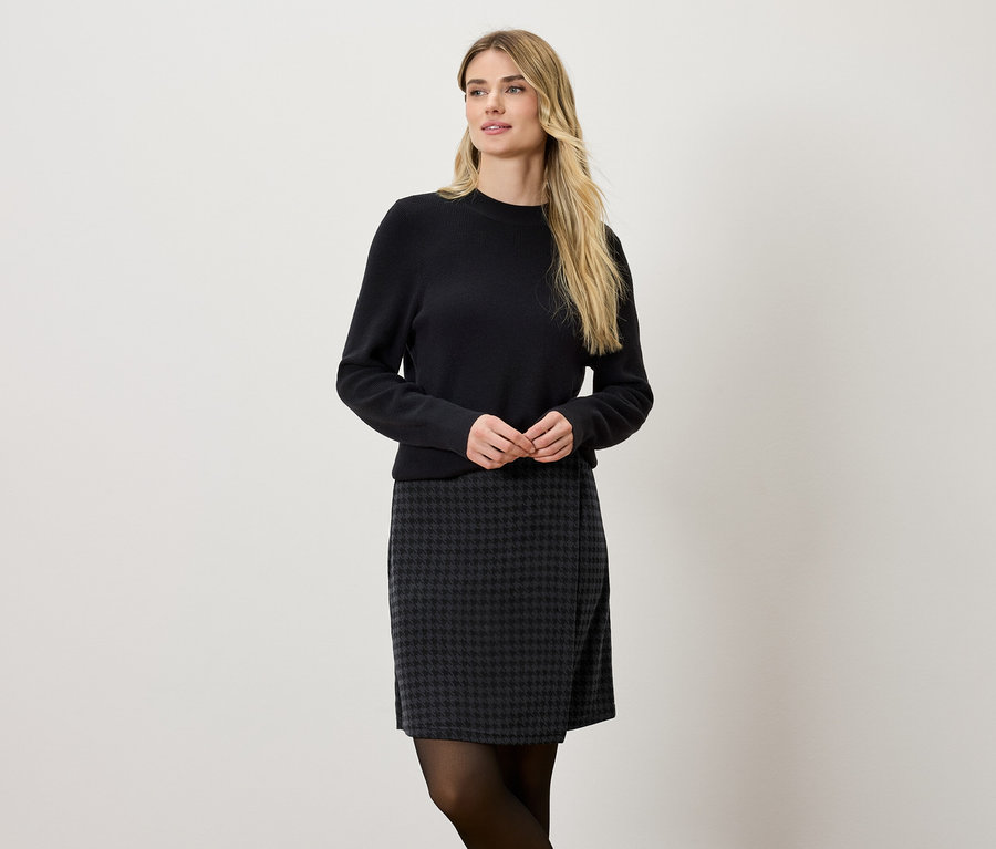 Blonde Frau trägt einen schwarzen Pullover und einen grau-schwarzen Rock.