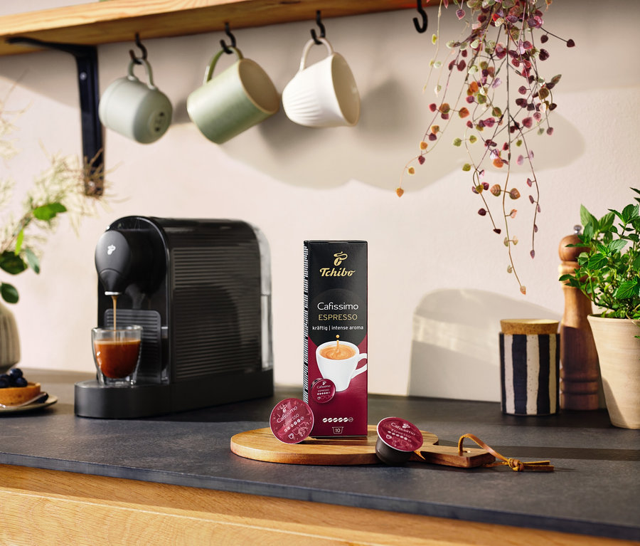 Eine schwarze Cafissimo pure+ bereitet Kaffee in ein Glas zu. Auf dem Tisch liegen eine Packung Kapseln und zwei Kapseln.