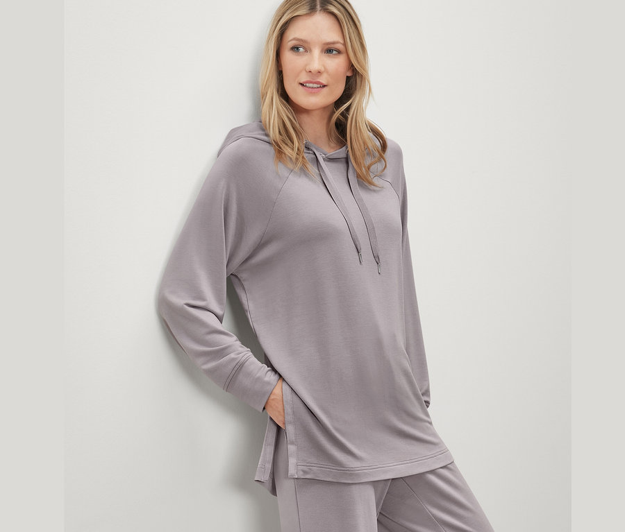 Frau in grauem Set mit Lounge-Hoodie lehnt an der Wand.