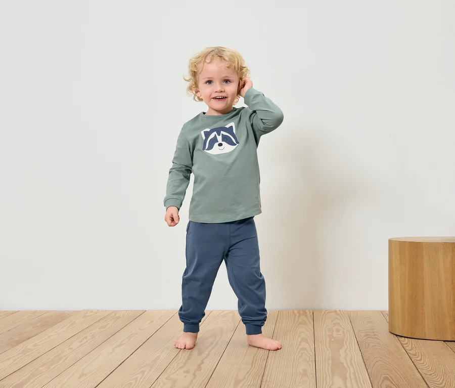 Ein blonder Junge in einem grünen Waschbär-T-Shirt und blauen Hosen steht barfuß auf einem Holzboden.