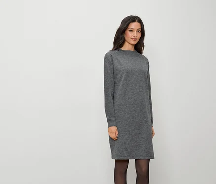 Eine Frau in einem langen, grauen Kleid und schwarzer Strumpfhose steht vor einem weißen Hintergrund.