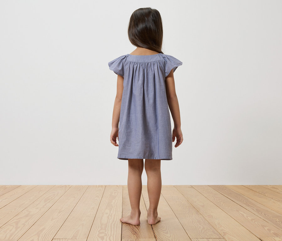 Rückansicht eines jungen Mädchens mit dunklen Haaren, das ein blaues Kleid trägt und barfuß auf einem Holzboden vor einer weißen Wand steht.