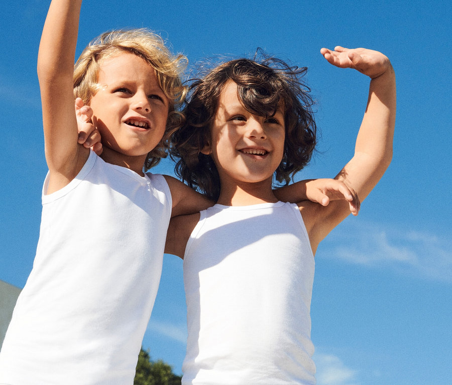 Zwei lachende Kinder mit blonden und braunen lockigen Haaren umarmen sich vor einem strahlend blauen Himmel und tragen weiße Tanktops.