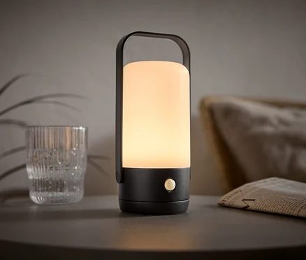 Eine beleuchtete, tragbare Lampe mit einem schwarzen Griff steht auf einem Tisch neben einem Glas und einer Zeitung.
