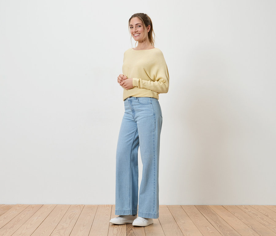 Frau mit Pferdeschwanz, gelbem Pullover, hellblauen Jeans und weißen Schuhen steht vor einer weißen Wand auf Holzboden und lächelt.