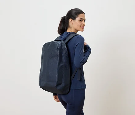 Frau mit marineblauem Trainingsanzug und marineblauem Rucksack, der über ihre Schulter hängt, blickt über ihre Schulter.