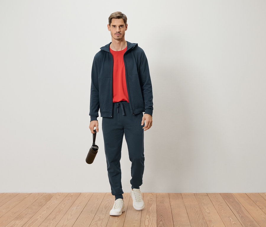 Mann steht in marineblauem Trainingsanzug und rotem T-Shirt, hält eine Trinkflasche in der Hand.