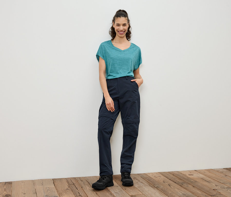 Eine Frau steht vor einer weißen Wand und trägt ein blaugrünes T-Shirt, eine blaue Zip-Off-Hose und schwarze Schuhe.