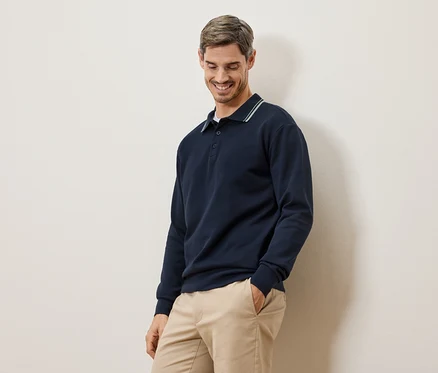 Ein lächelnder Mann in einem marineblauen Langarmpolohemd und beigen Chinos steht vor einer cremefarbenen Wand.