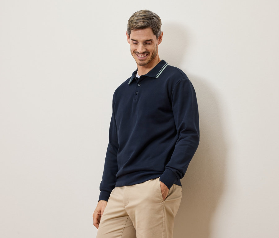 Ein lächelnder Mann in einem marineblauen Langarmpolohemd und beigen Chinos steht vor einer cremefarbenen Wand.